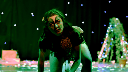 A performing arts student in green costume and face paint, is on their knees with a blank but stern facial expression