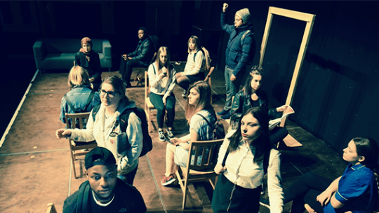 12 Performing arts students sat at desks and some stood in a school style performance