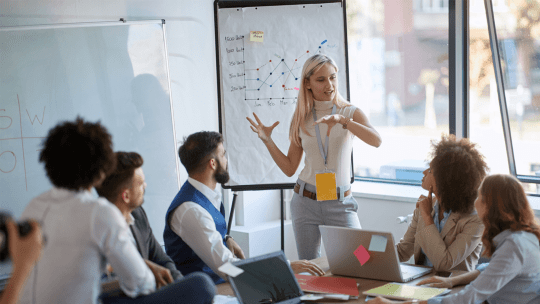 A woman stood in front of a white board talking to 5 adults all sat at a table looking in her direction
