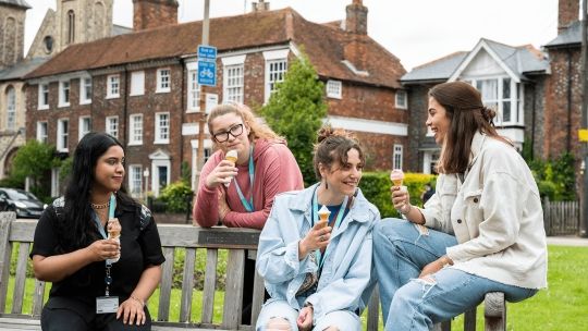 Four female BNU students sat on a bench each with an ice cream in their hand looking at one another and smiling in High Wycombe town centre
