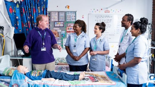 Four student nurses in uniform stood around a simulation hospital bed with a child laying down on it