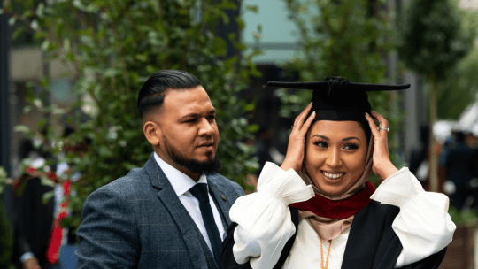 A graduate rearranging her hat whilst a man stood next to her looks at her in admiration
