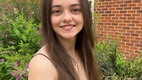 Student Sophie, who has pale skin and waist-length brown hair, stands in front of a brick building.