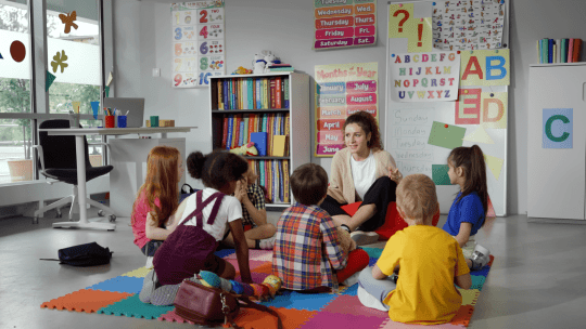 Six pre-school children sat on the floor of a classroom looking at their teacher. 