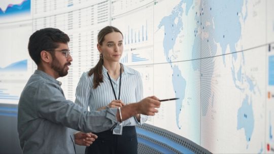 two people talking in front of a digital screen with a map, graphs and charts