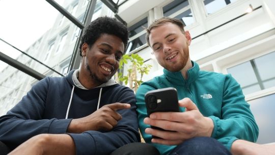 Two smiling male students sitting together in a modern, bright atrium with a glass ceiling, looking at a smartphone held by the student in the teal fleece.