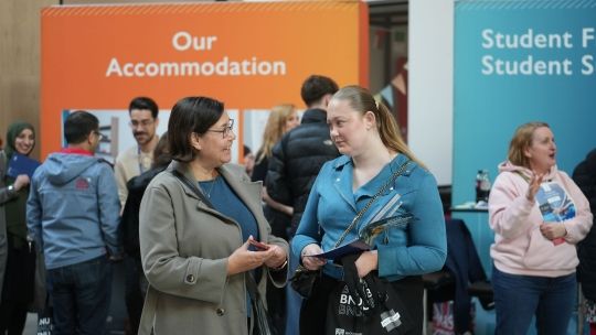 A mother and daughter talk at a BNU Open Day, standing in front of information stands for student accommodation and finance while holding university brochures.