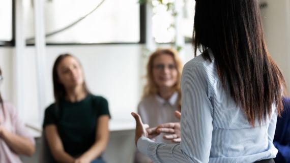 woman standing up talking to two others who are sat down on chairs