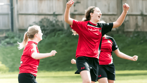 Three women from the BNU football team celebrating on the football pitch, wearing their red and blue kit