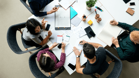 An aerial view of a table with 5 students sat around it looking and discussing at meeting notes 