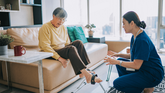 An occupational therapist kneeling down as a patient is sat on a sofa doing some foot exercises.