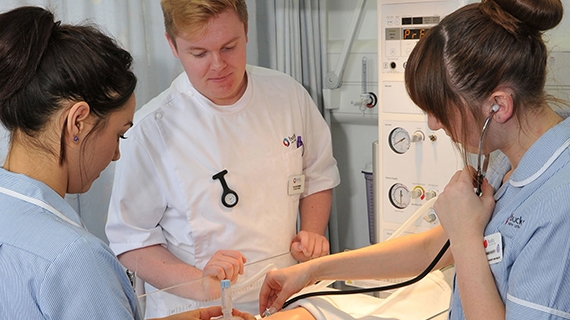 Three Nursing students stood around a bed