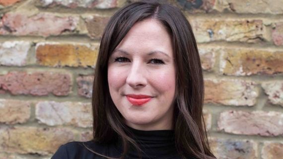 A head and shoulders shot of Lara Baker stood with her arms crossed wearing a black jumper and looking directly into the camera
