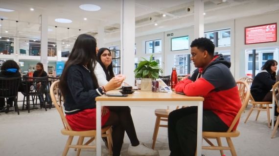 Three students sat down at a table in the canteen chatting and eating food.