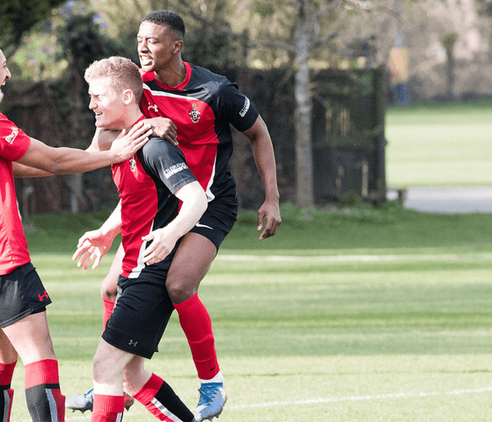 Three BNU footabllers celebrate scoring a goal. One players gives another a piggy back and then goes to hug the third person