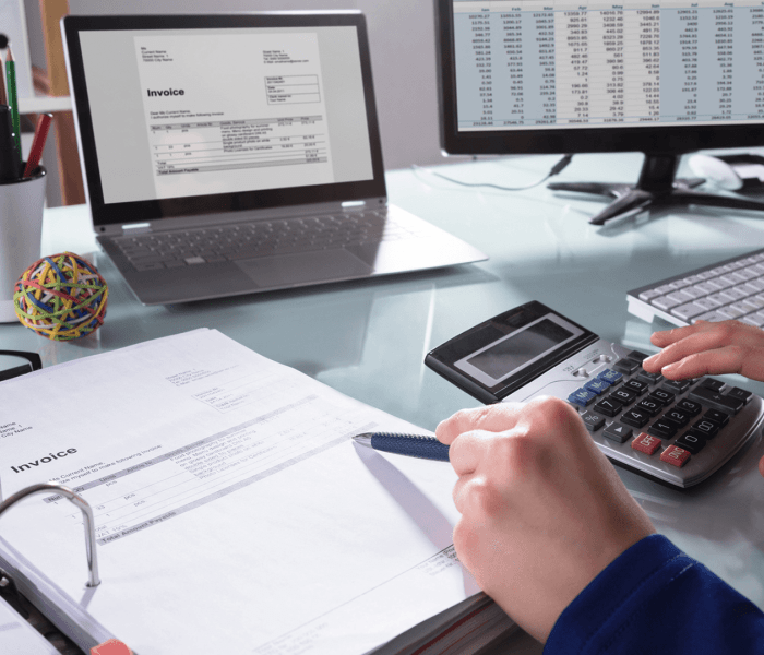 An accounting and finance student sat at a desk looking at invoice information on two computer screens with a calculator