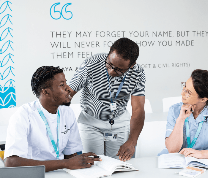 Two student nurses sat at a desk and a lecturer stood looking at the students talking through their work 