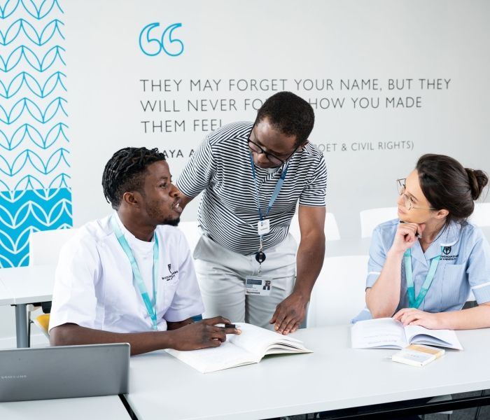 Two nursing students sat at a desk in a class with the teacher stood in between them in discussion