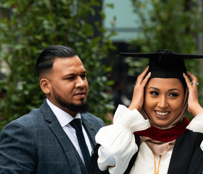 A graduate rearranging her hat whilst a man stood next to her looks at her in admiration