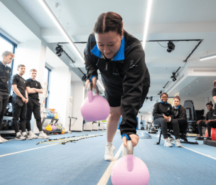 A Sports Therapy student on the indoor running track leaning down to pick up a weight from the floor.