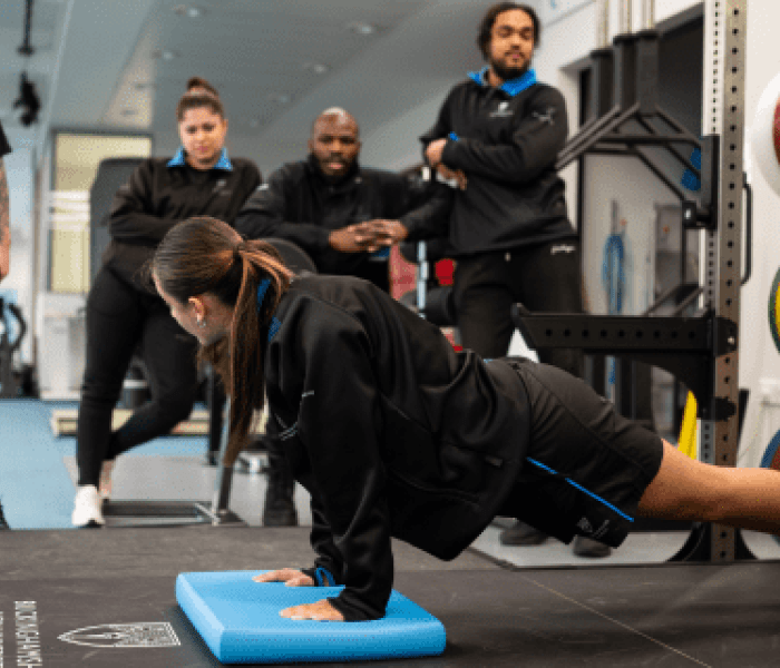 A Sports Therapy student doing a press up on the floor as four other students watch on.