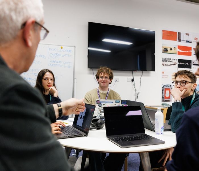 Students sitting around a table with their laptops, talking to a lecturer
