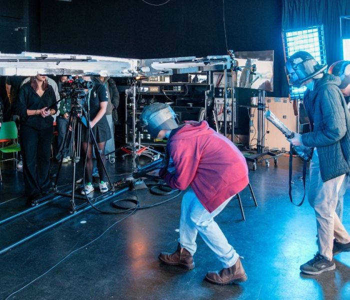 Three students acting on set whilst a group of Film students control the lighting and cameras behind scene.