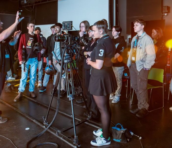 A group of students stood around a lecturer who has their hand raised whilst talking to the group in a Film and TV studio.