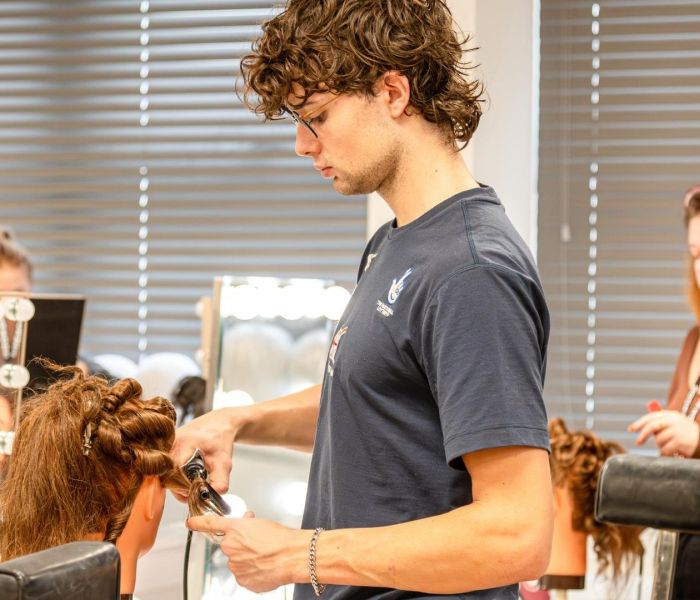 A Hair and make-up student stood up working on a wig in a classroom, with a fellow student in the back of the room..