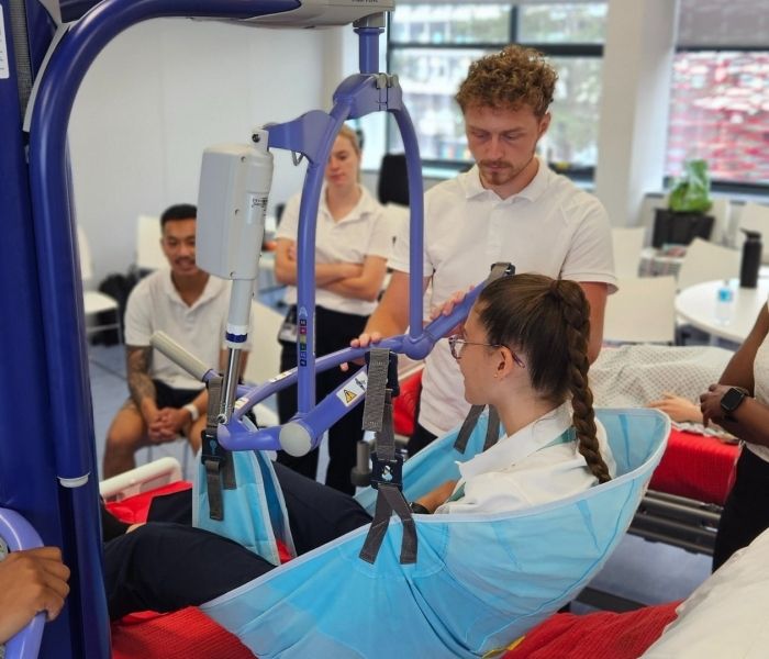 Five Physiotherapy students all in white t-shirts stoof around a lecturer talking to a student laying above a bed in a hoist.