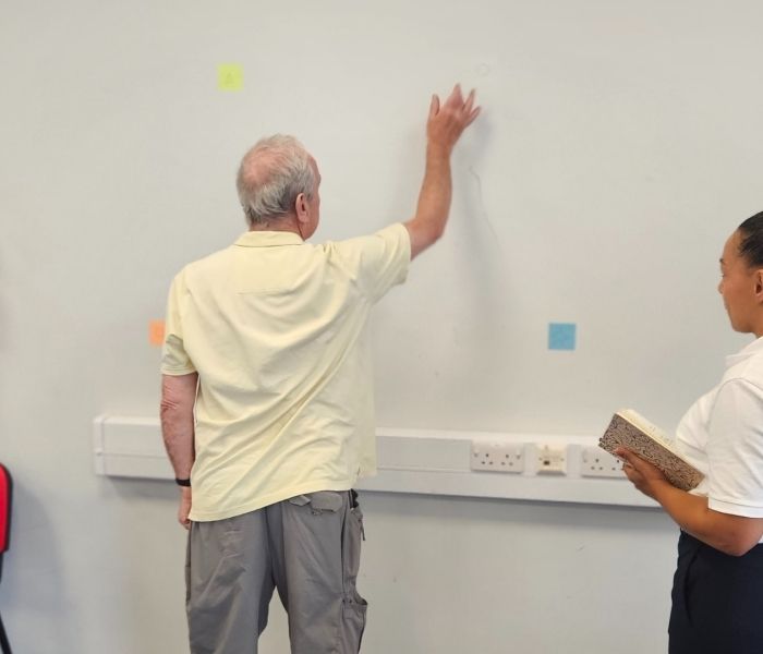 An elderly man raises his right hand against a wall as two Physiotherapy students watch on.