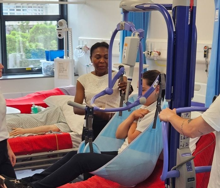 Three Physiotherapy students stood around a hospital simulation bed as a fellow student is sat in a hoist above a bed.