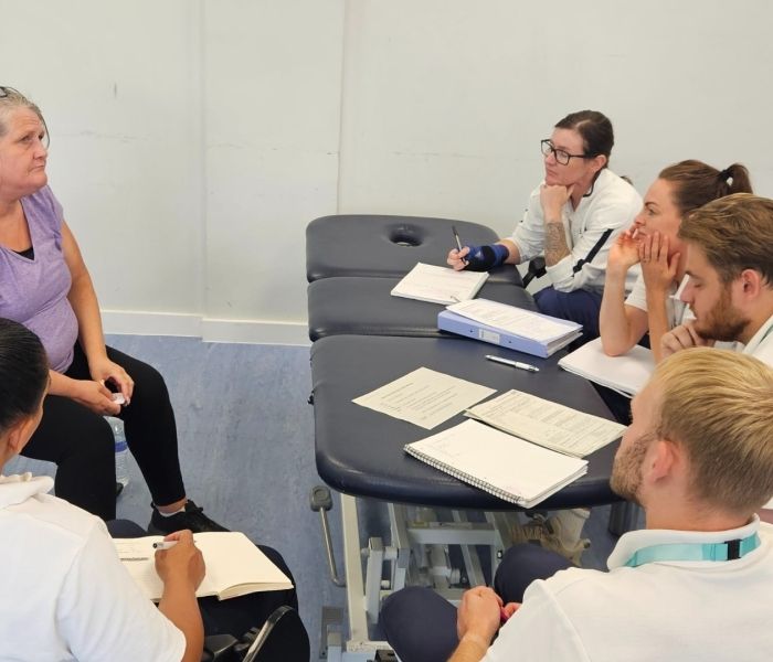 A member of the public sat on a chair in front of five Physiotherapy students with notebooks laid on a table.