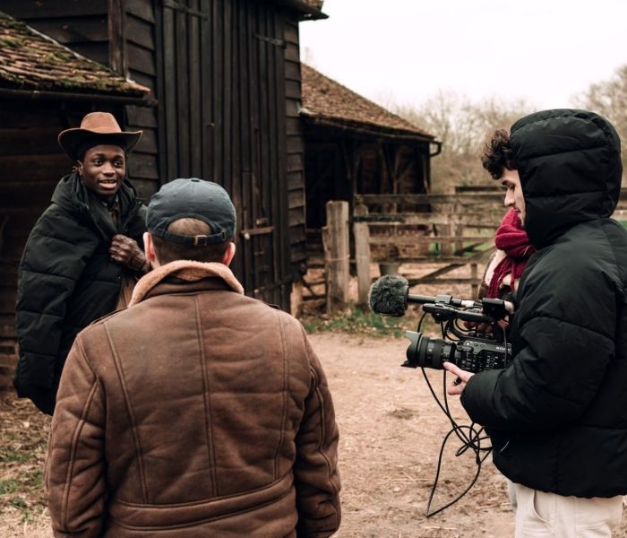 A cameraman wearing a coat stood next to another student as they both face a man wearing a cowboy cap in a stables setting.