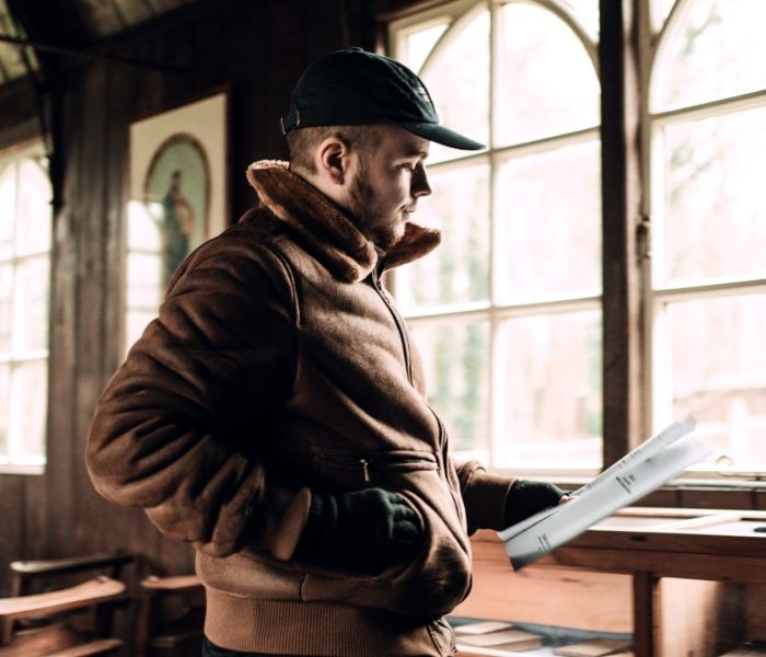 A student stood in an old buliding wearing a cap and holding a script in their left hand.