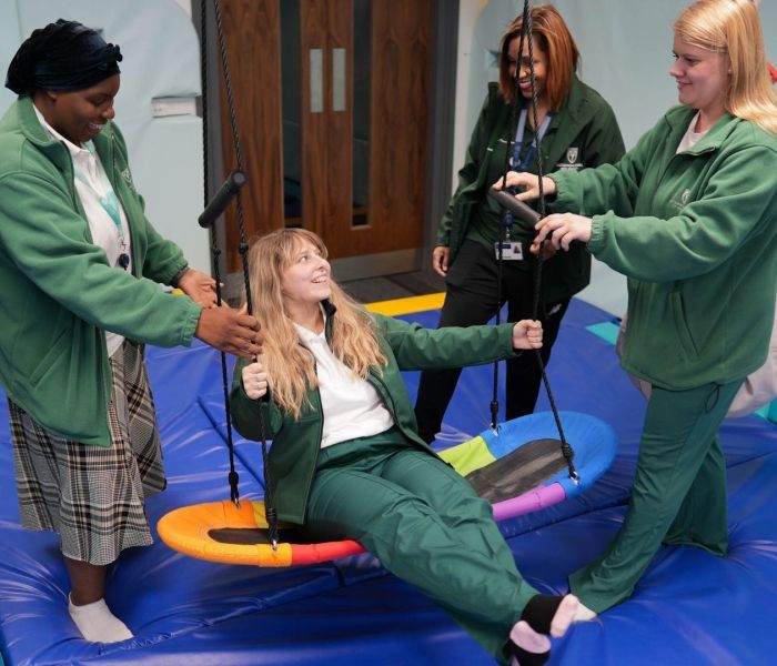 Three Occupational Therapy students stood either side of an indoor swing as another student holds onto the handles and swings.