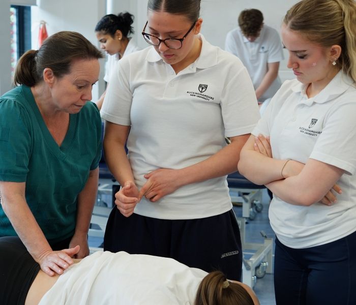 Two Physiotherapy students stood up observing a lecturer giving a massage on the lower back of a fellow student in a classroom setting.