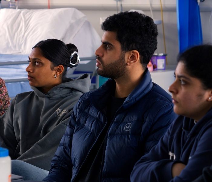 Three Physiotherapy students sat next to one another in a fake hospital setting with a concentrated look on their faces.