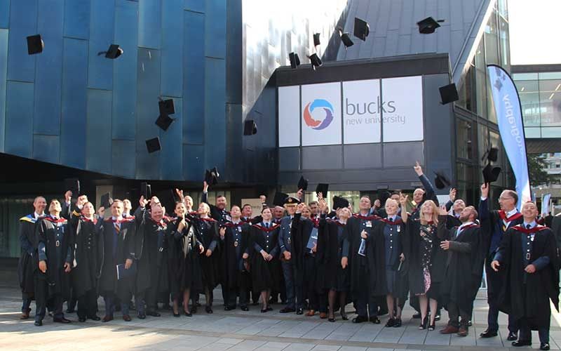 A large group of Organisational Resilience graduates throw up their graduation hats stood outside the gateway building