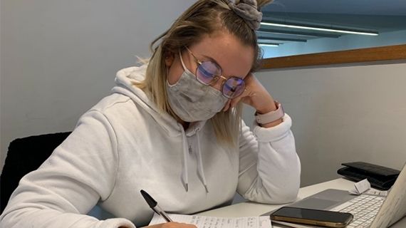 Student wearing glasses and a mask sat at a desk with a pen in her hand