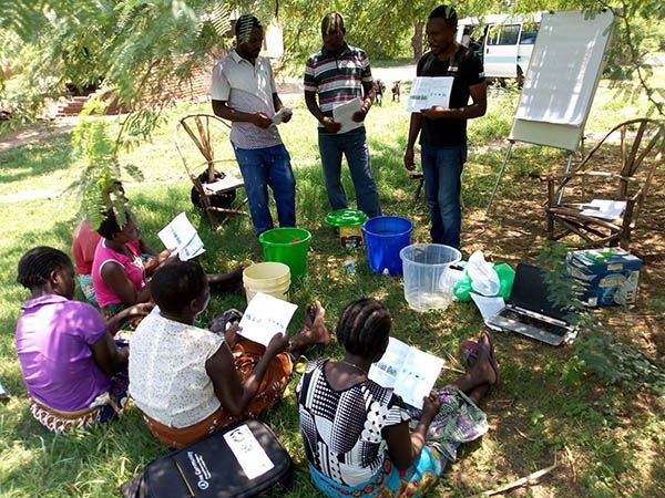 Three men stood up holding a bit of paper and five people sat down facing towards them on the grass