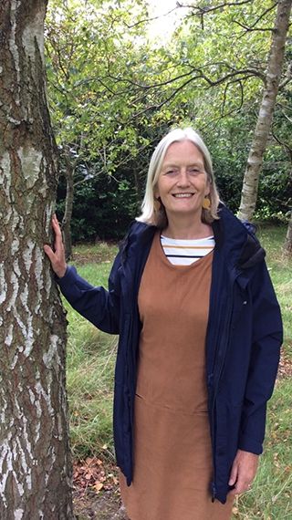 Helena Chance stood with her right hand leaning on a tree wearing a cardigan with a dress looking towards the camera