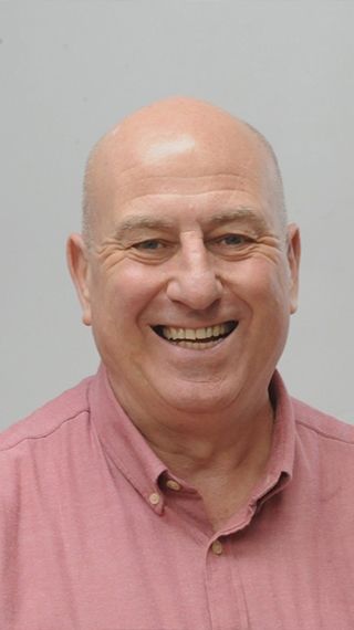 Head and shoulders shot of a smiling Richard Jones wearing a pink shirt stood in front of a grey wall looking into the camera