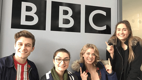 Four Performing Arts students sat below a BBC sign, with two students pointing up to it