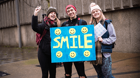 Three BNU students stood outside holding a banner with the word 'smile' written on it. 