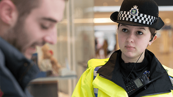 A police officer wearing a police hat and yellow high vis jacket, looking at a man who is looking down and smiling