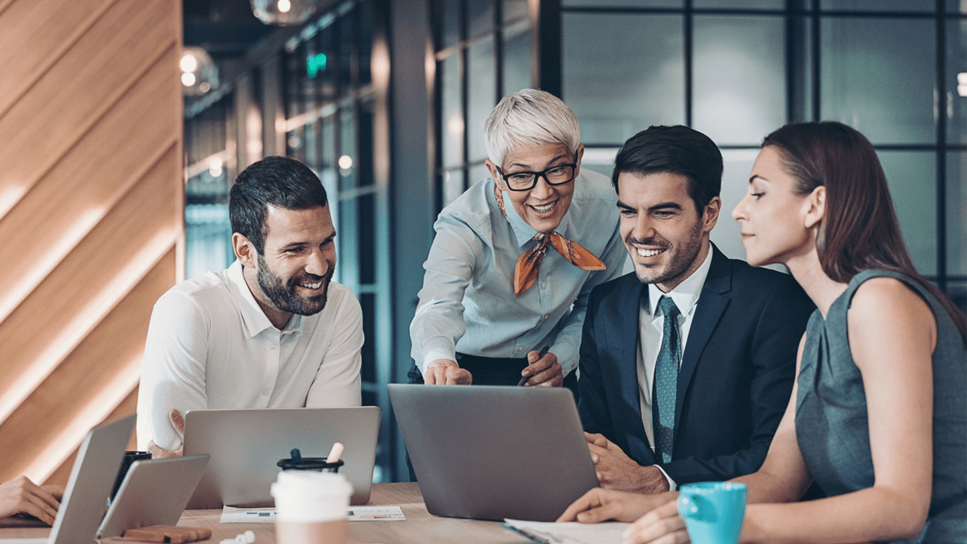 Three people sat looking around a laptop whist a woman is standing over looking at the laptop and smiling