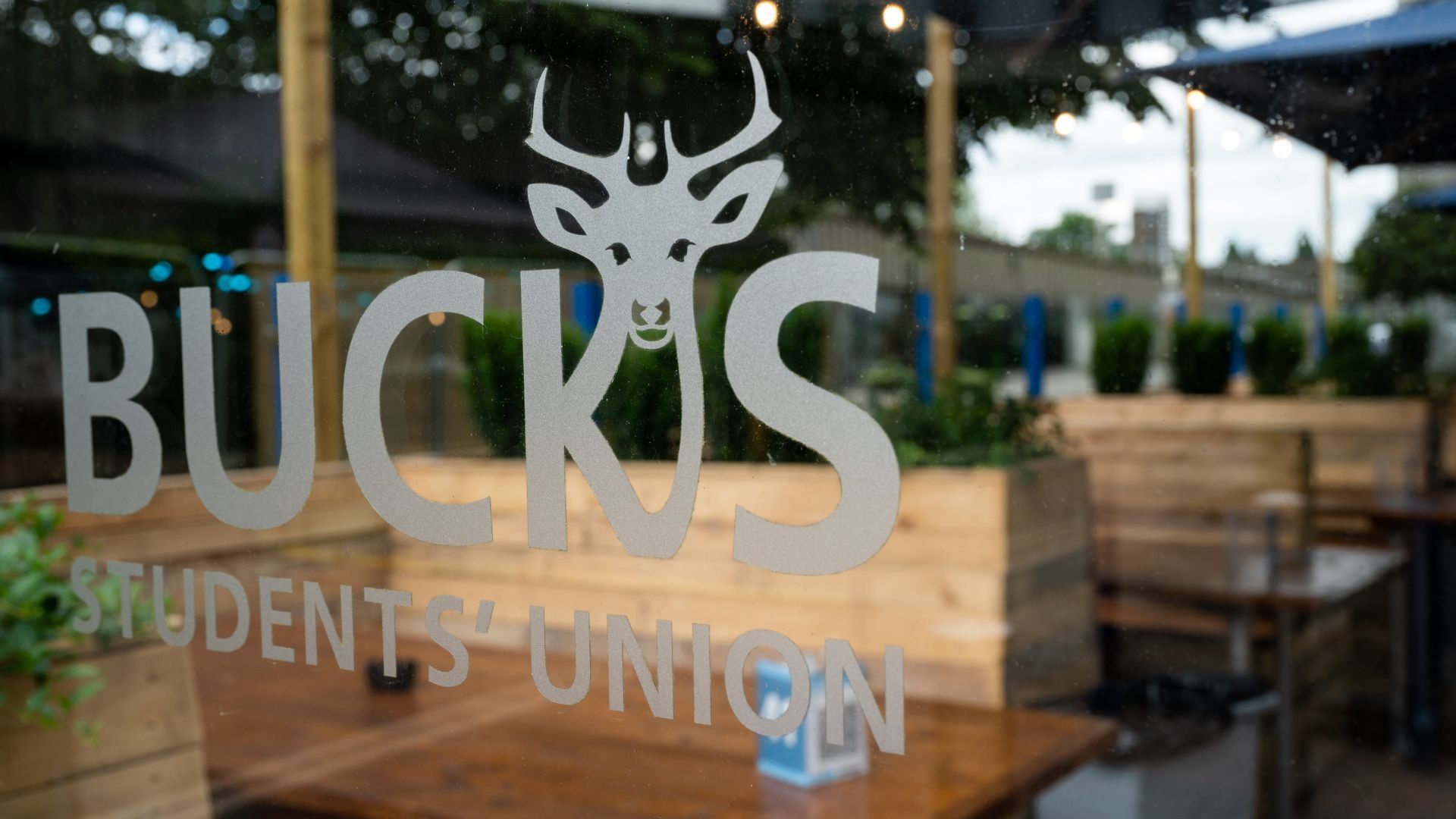 Bucks Students' Union logo on a pane of glass which has an outdoor wooden seating area behind it