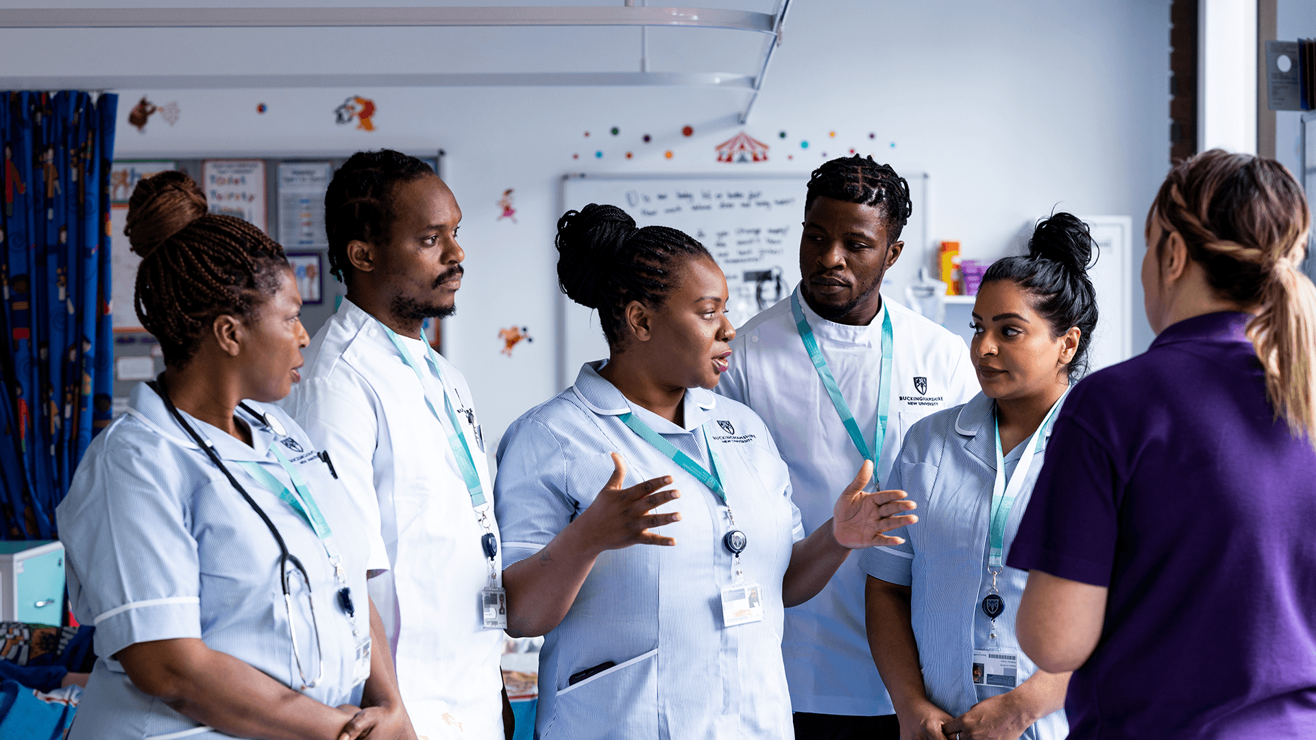 Five student nurses stood in conversation with a lecturer in a nursing simulation suite 