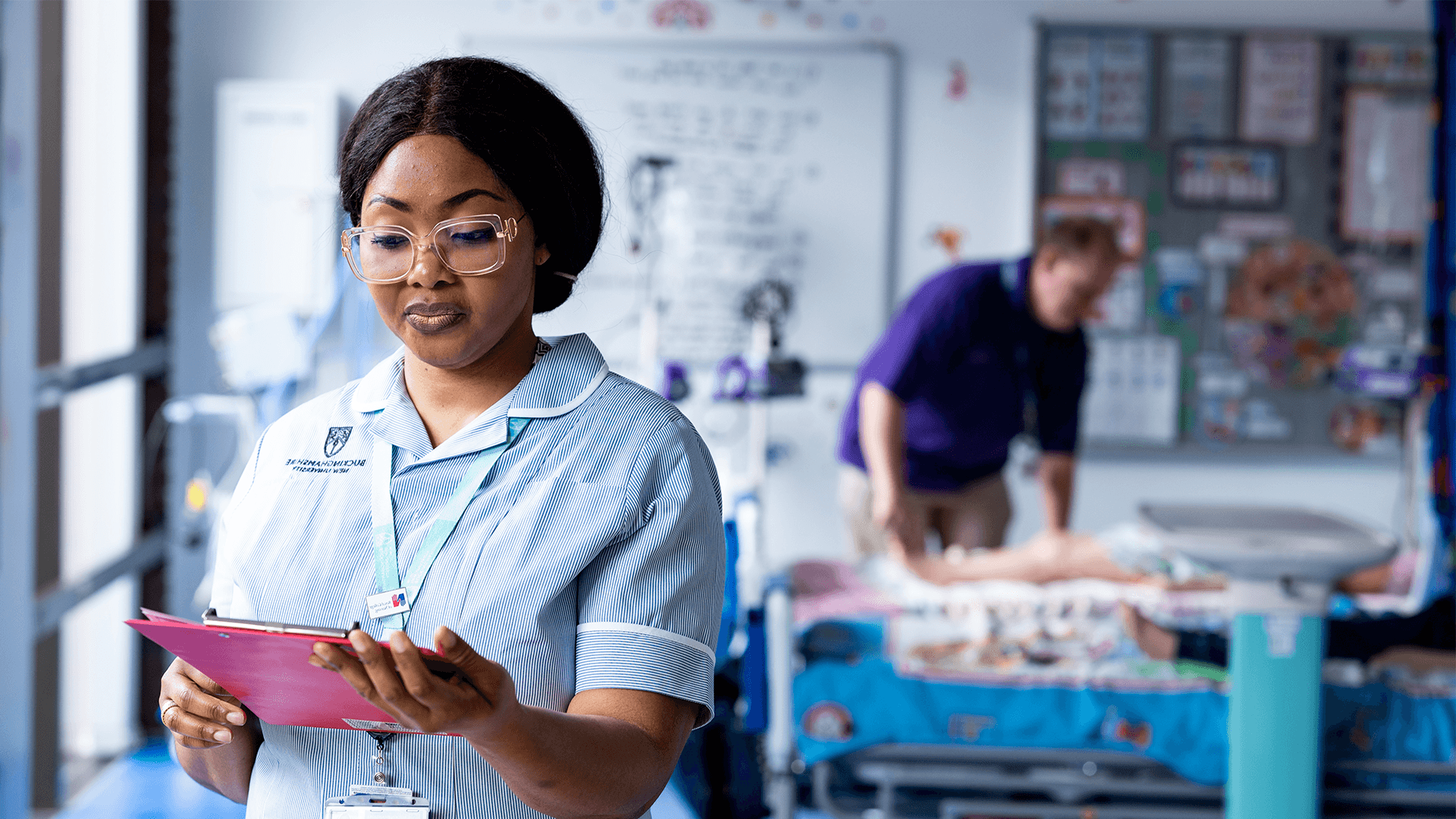 A female student nurse looking at her notes whilst a lecturer assists to a dummy patient in the background